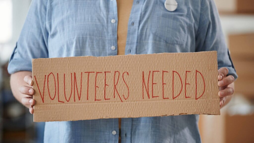 Person holding a sign that reads, "Volunteers Needed"